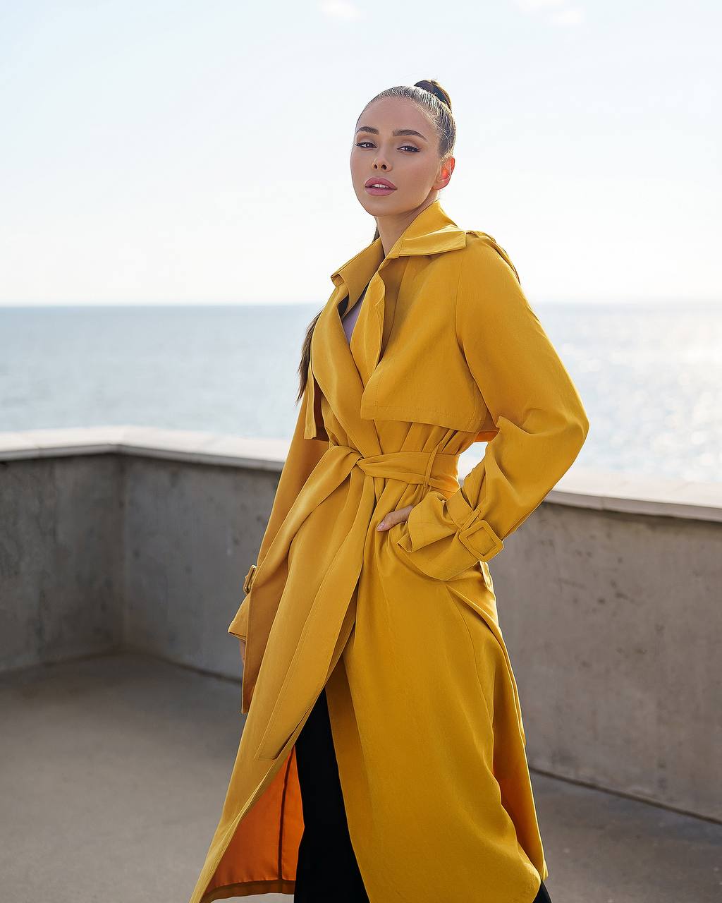 Woman wearing a yellow trench coat standing on a rooftop with ocean view