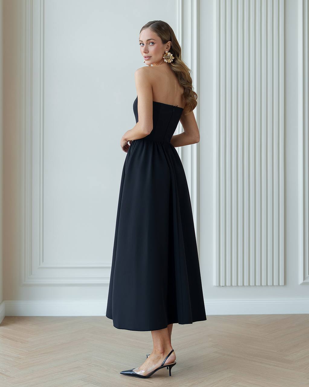 Woman wearing a black strapless dress standing against a white paneled wall.