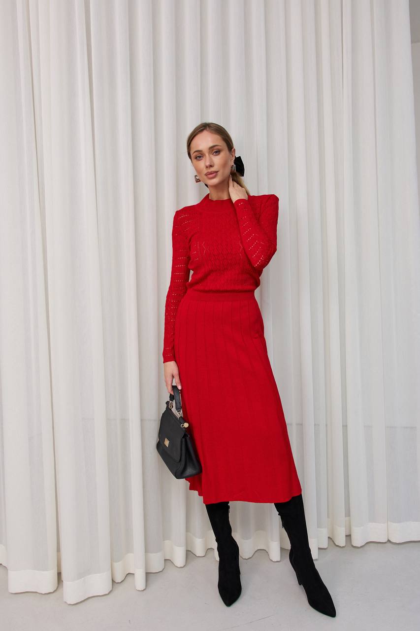 Woman wearing a red outfit with black boots and a black handbag against a white curtain background