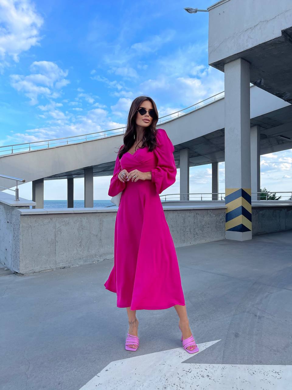 Woman in a bright pink dress standing on a rooftop with a blue sky and concrete structure in the background.