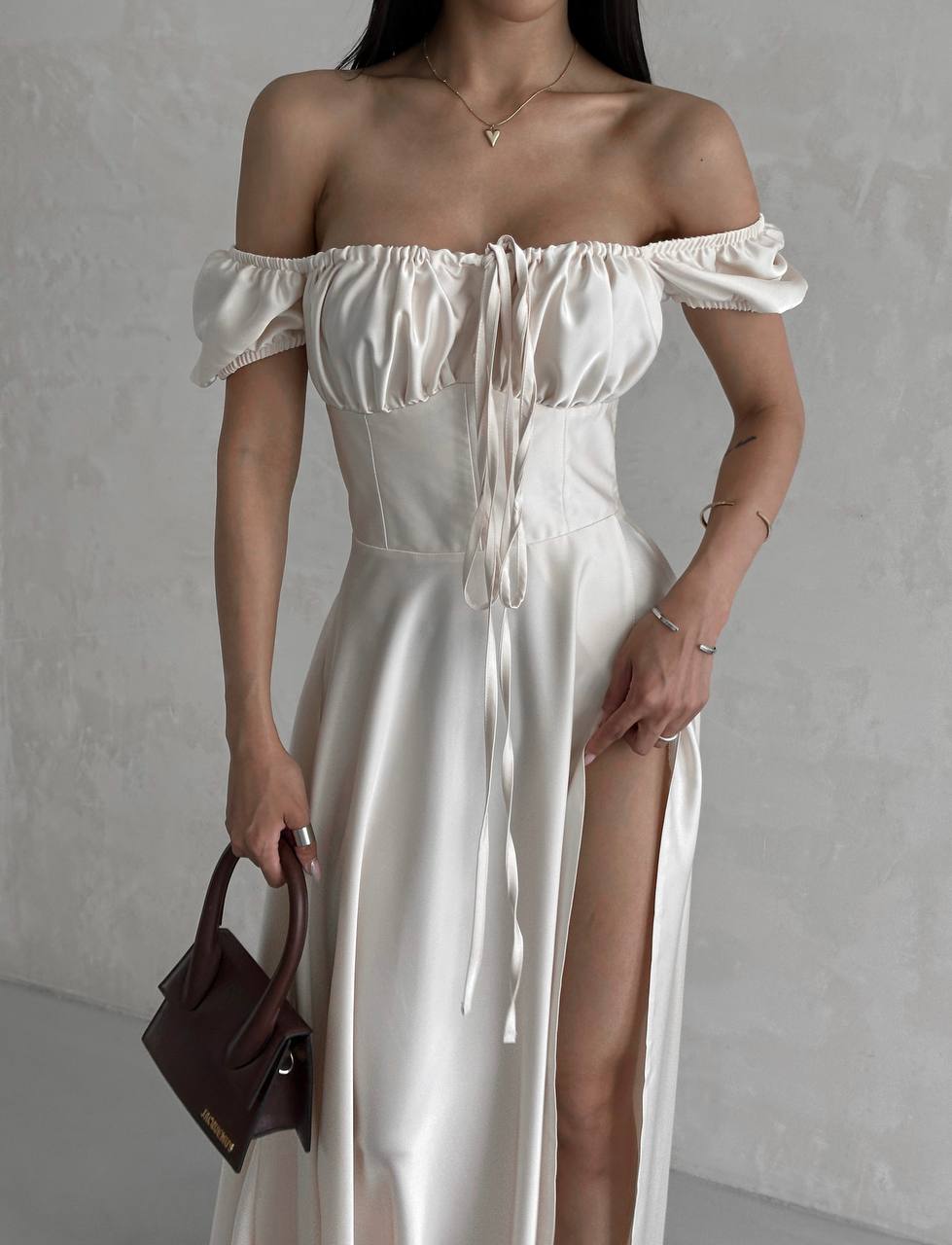 Woman wearing a white off-shoulder dress with a brown handbag against a plain background