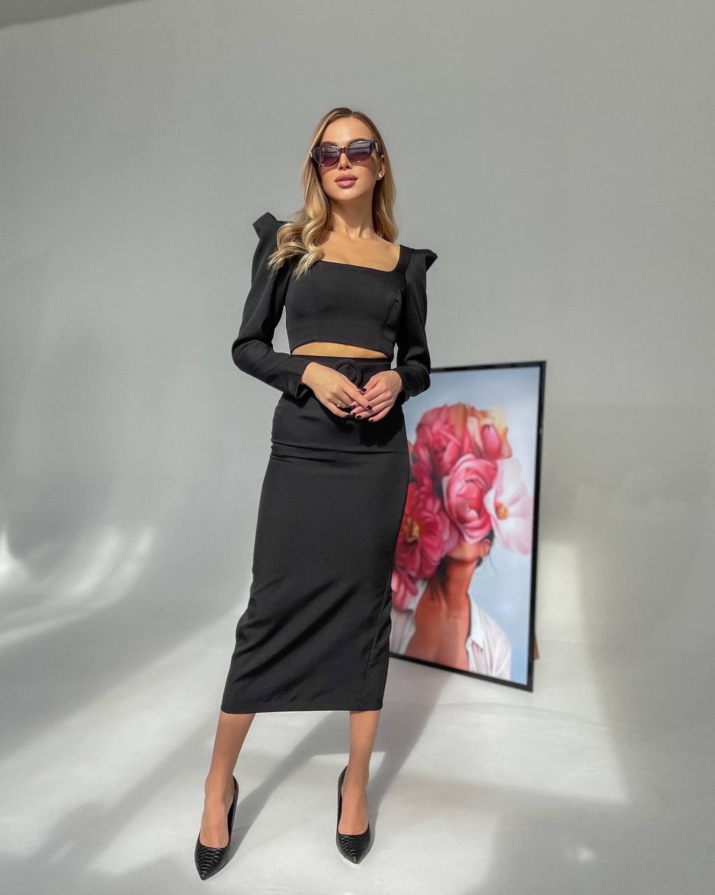 Woman in black outfit standing in front of a floral artwork on a gray background