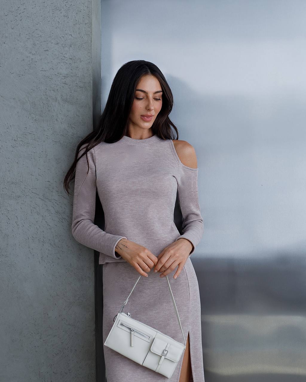 Woman in a gray dress holding a white handbag against a gray wall