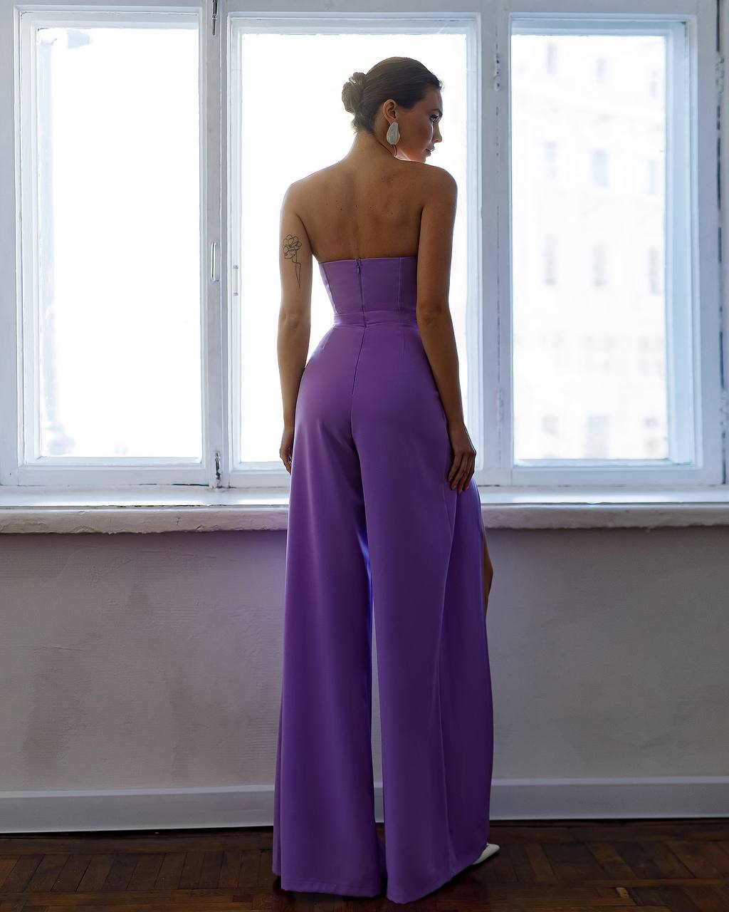 Woman wearing a purple strapless jumpsuit standing in front of a window.