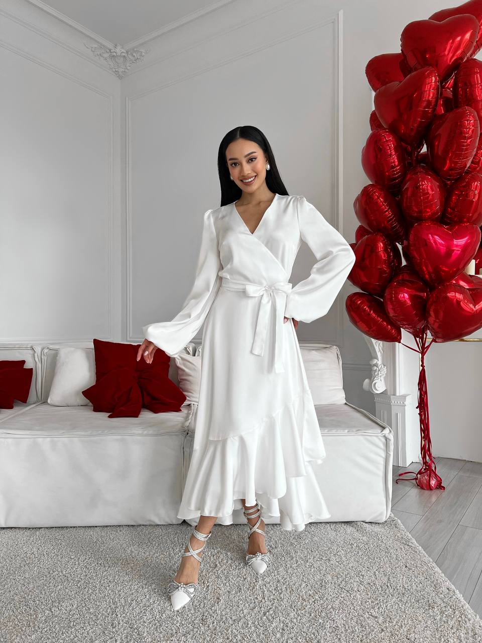 Woman in a white dress standing in a room with red heart-shaped balloons.