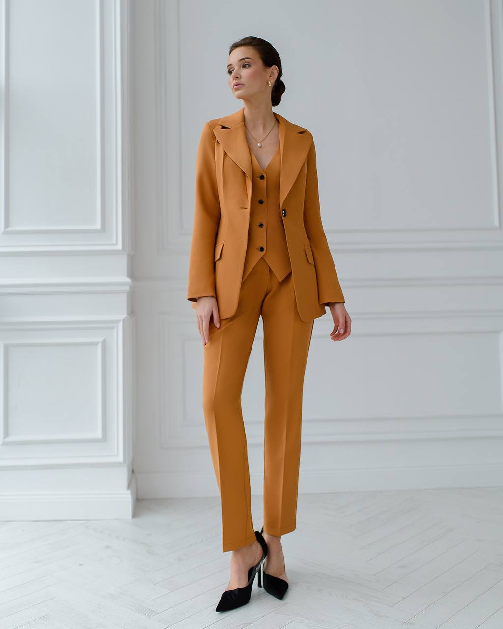 Woman wearing a mustard yellow suit standing against a white paneled wall.