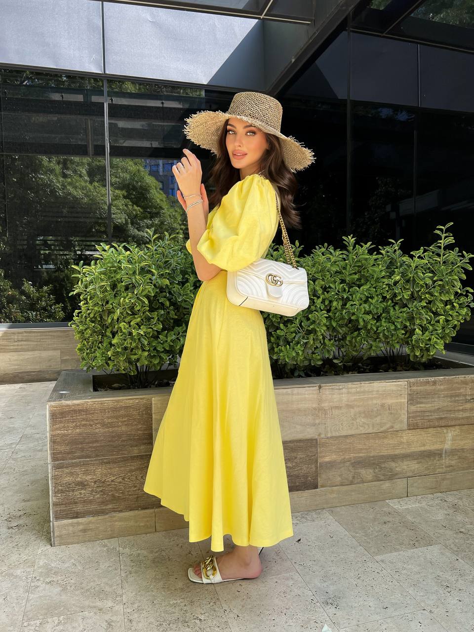 Woman in a yellow dress and straw hat standing outdoors with greenery and a building in the background.