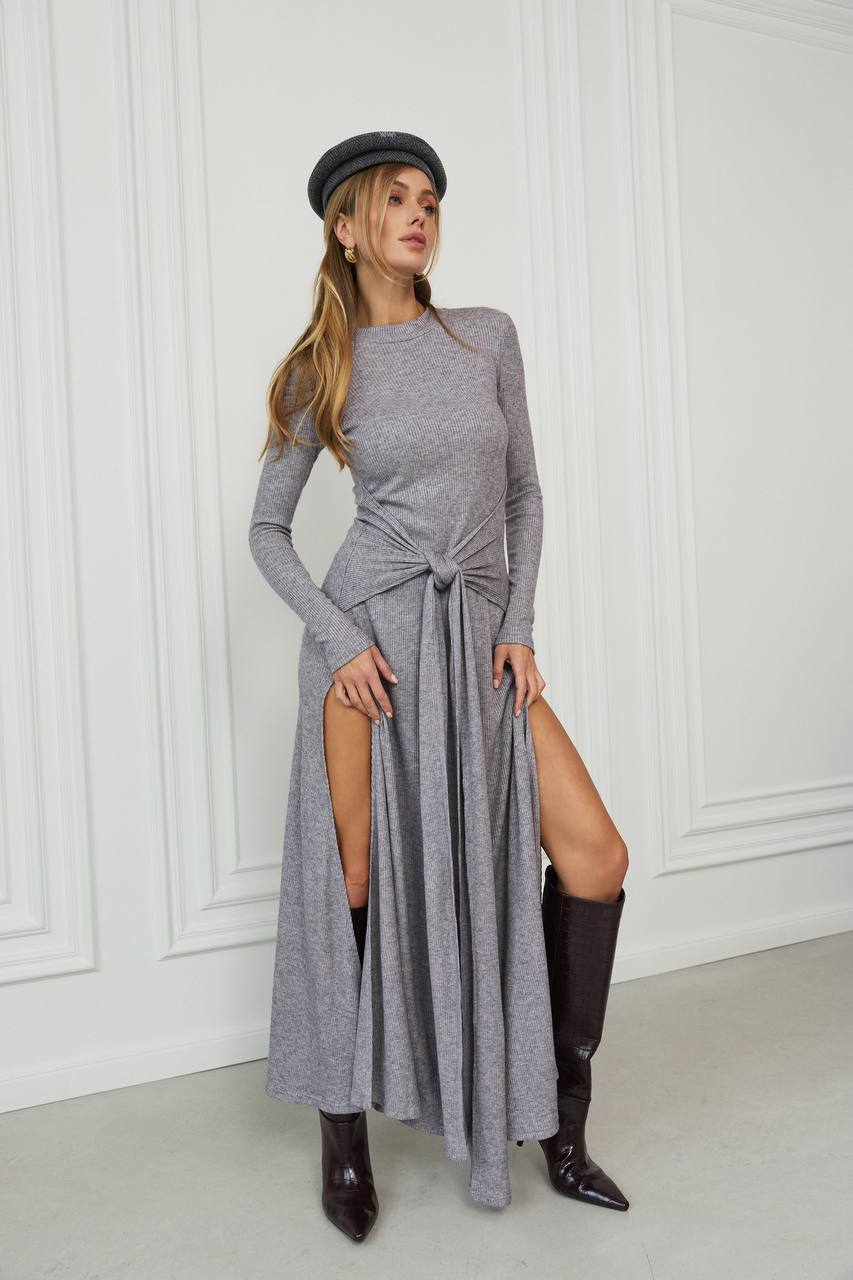 Woman wearing a long gray dress with a slit, black knee-high boots, and a black beret against a white paneled wall.