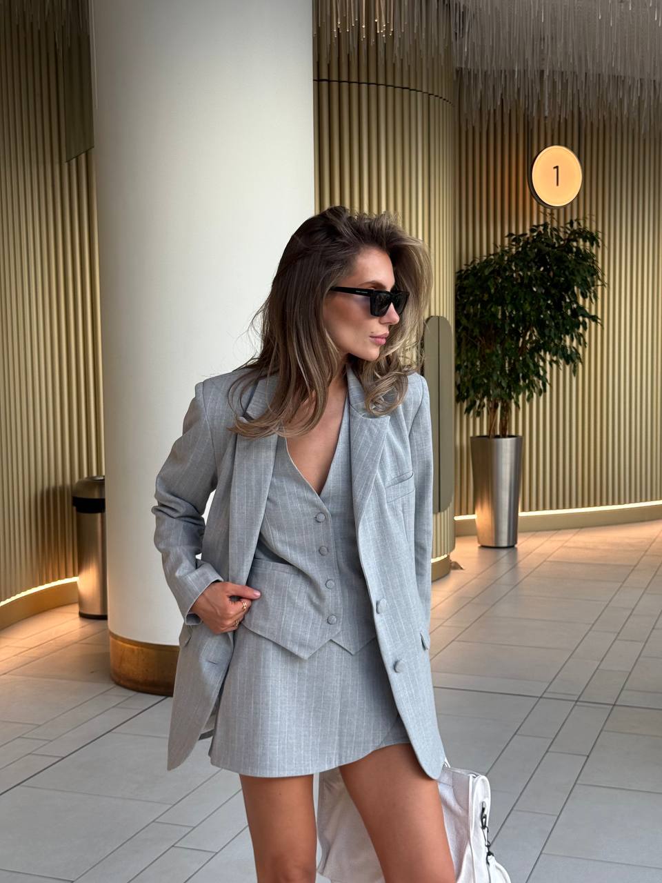 Woman in a gray suit standing in an indoor setting with a plant and light fixture in the background.