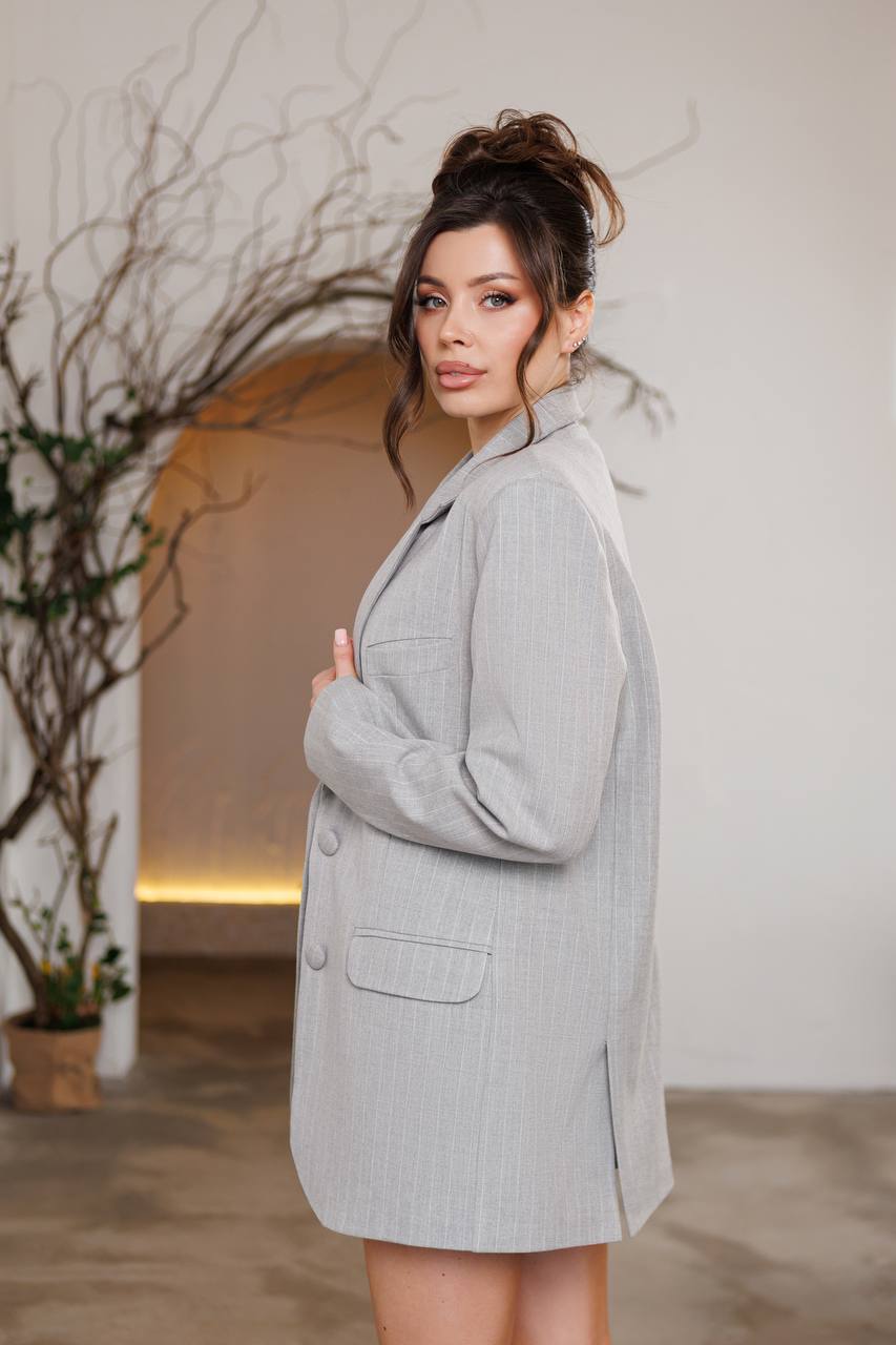 Woman wearing a gray coat standing in a softly lit room with a plant in the background