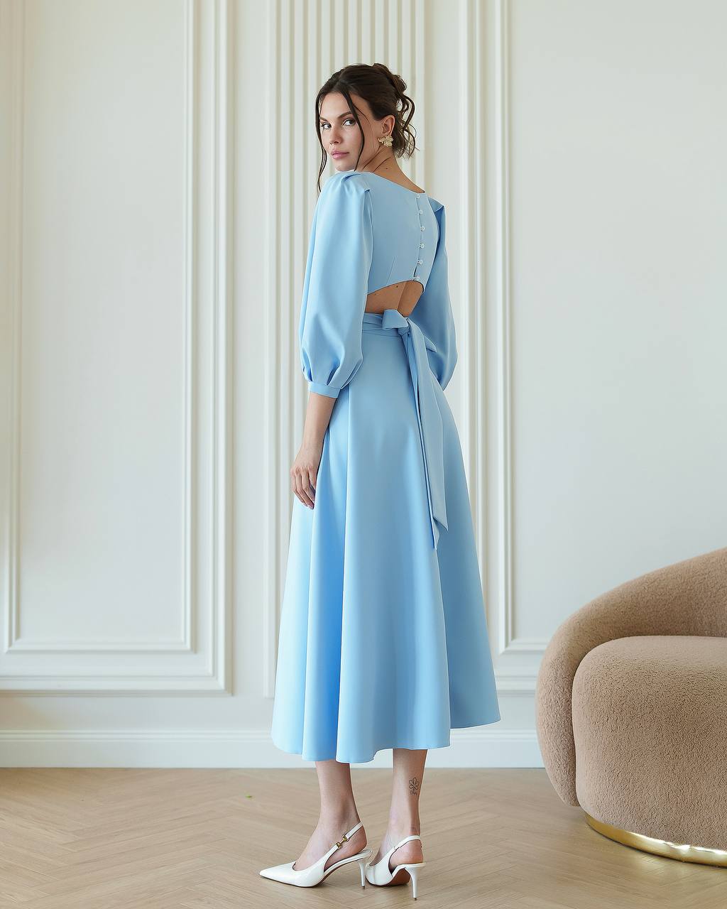 Woman wearing a light blue dress standing in a room with white walls and a beige chair.