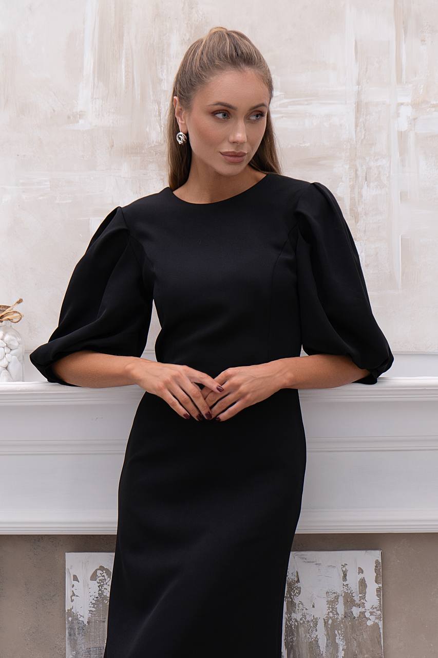 Woman wearing a black dress with puffed sleeves against a white wall.
