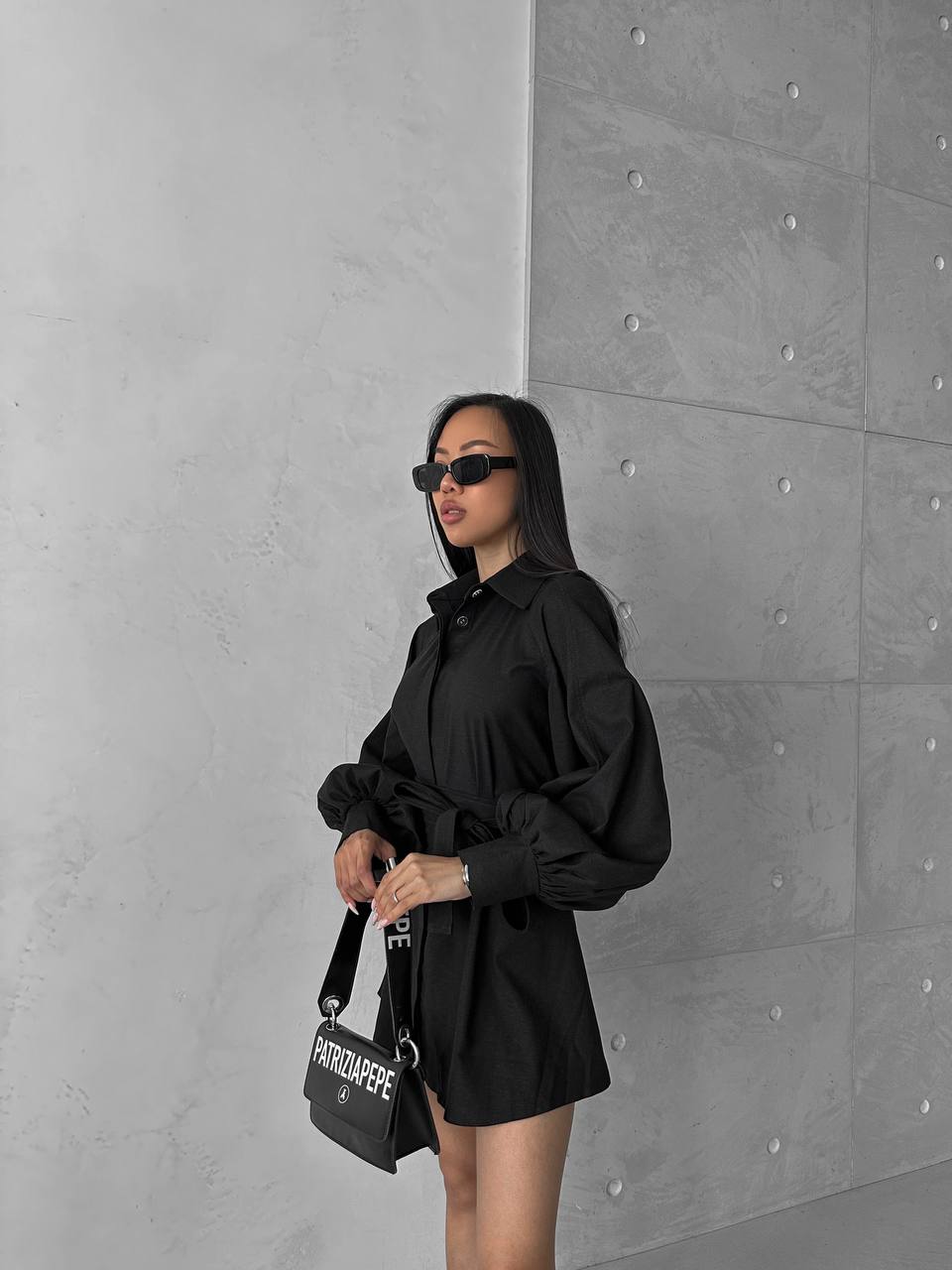 Woman in a black outfit holding a black handbag against a gray concrete wall.