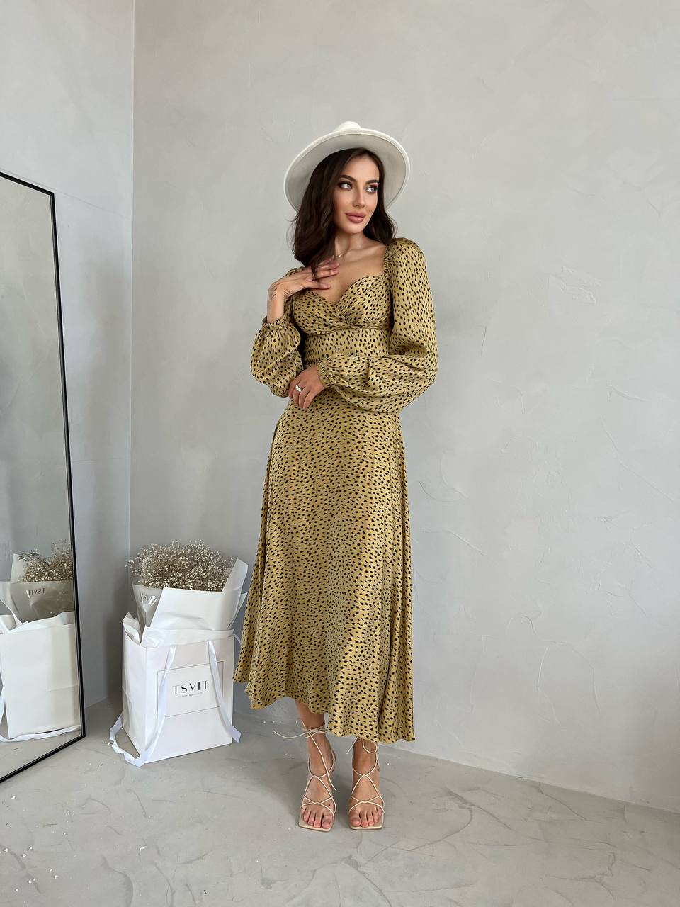 Woman wearing a gold patterned dress and white hat in a minimalistic room.