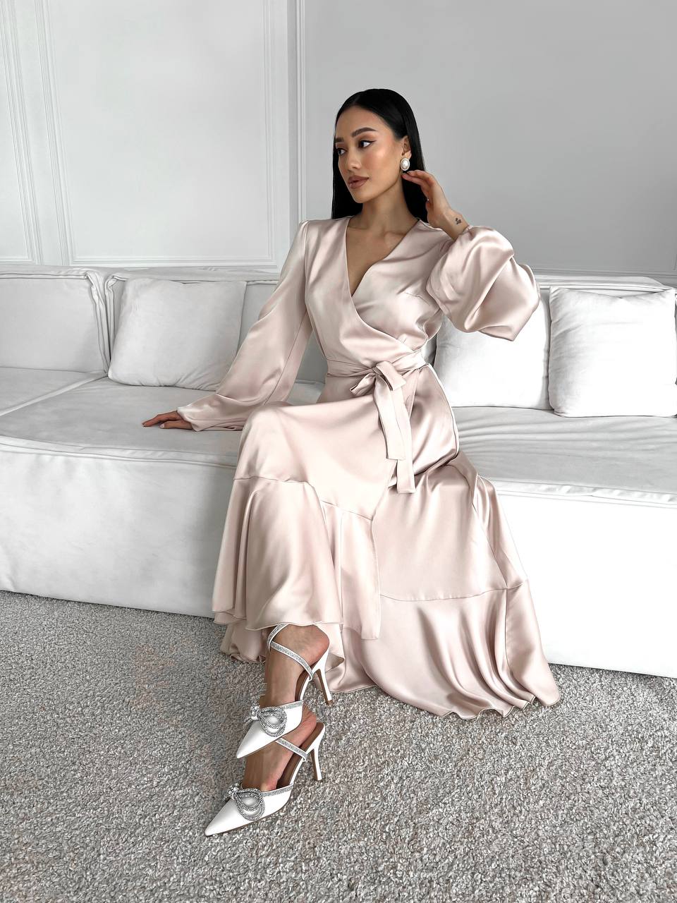 Woman in a light pink satin dress sitting on a white couch in a minimalistic room.