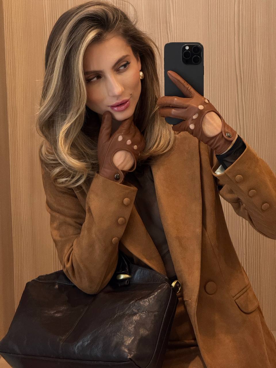 Woman in a brown coat and gloves taking a selfie against a wooden wall.