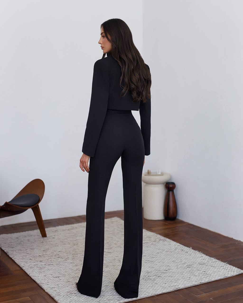 Woman in a black suit standing in a minimalistic room with a white wall and wooden floor.