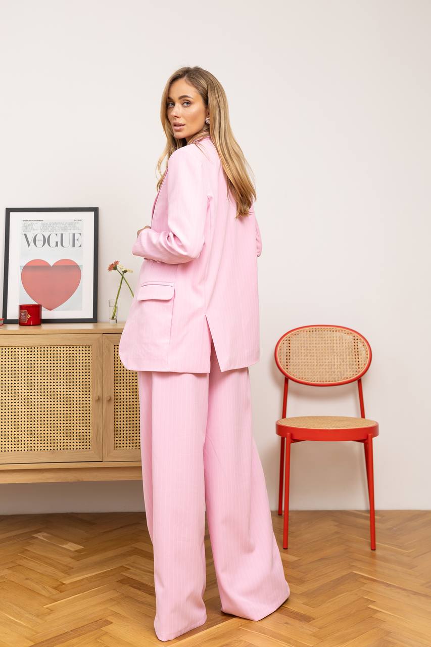 Woman in a pink suit standing in a room with a red chair and wooden cabinet.