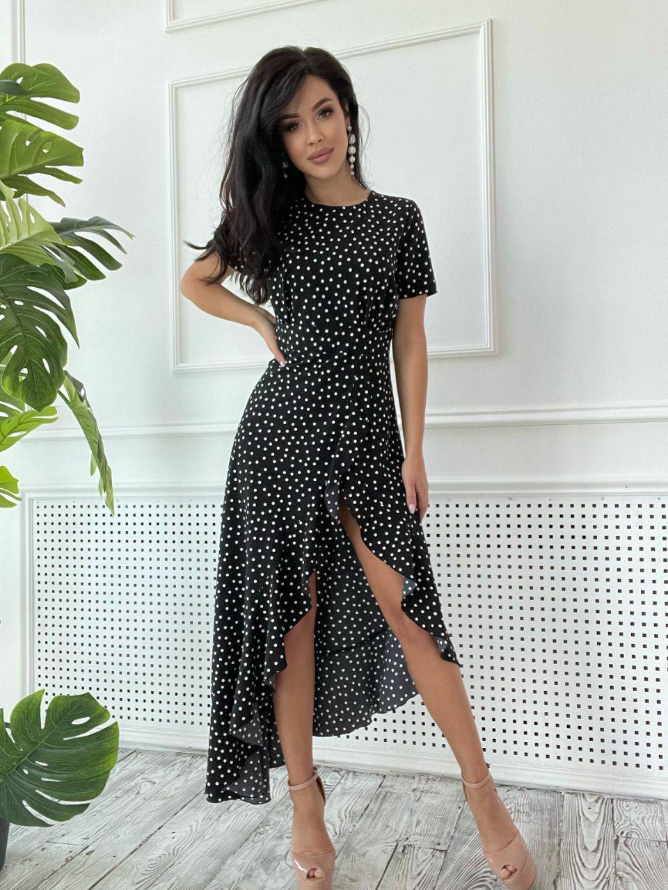 Woman wearing a black polka dot dress in a room with plants and a white wall.