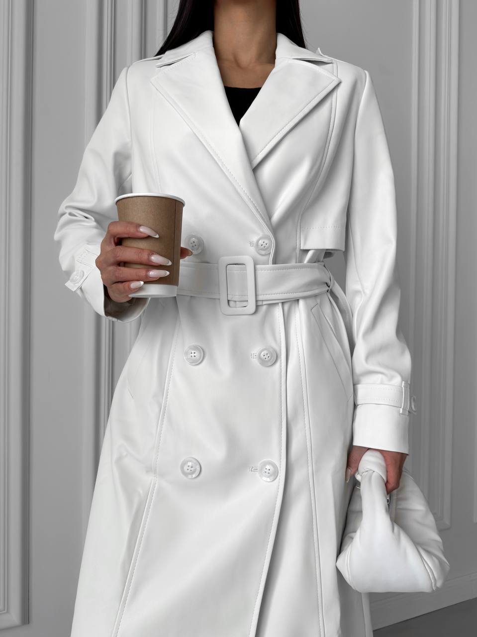 Person wearing a white trench coat holding a coffee cup against a gray wall.