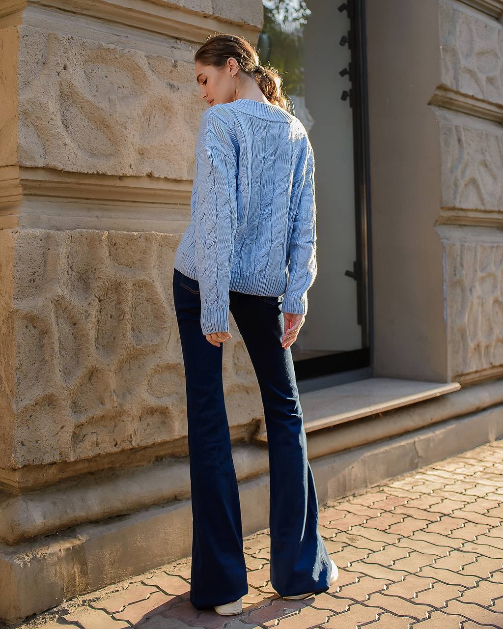 Woman wearing a light blue cable knit sweater and dark blue jeans standing against a stone wall.