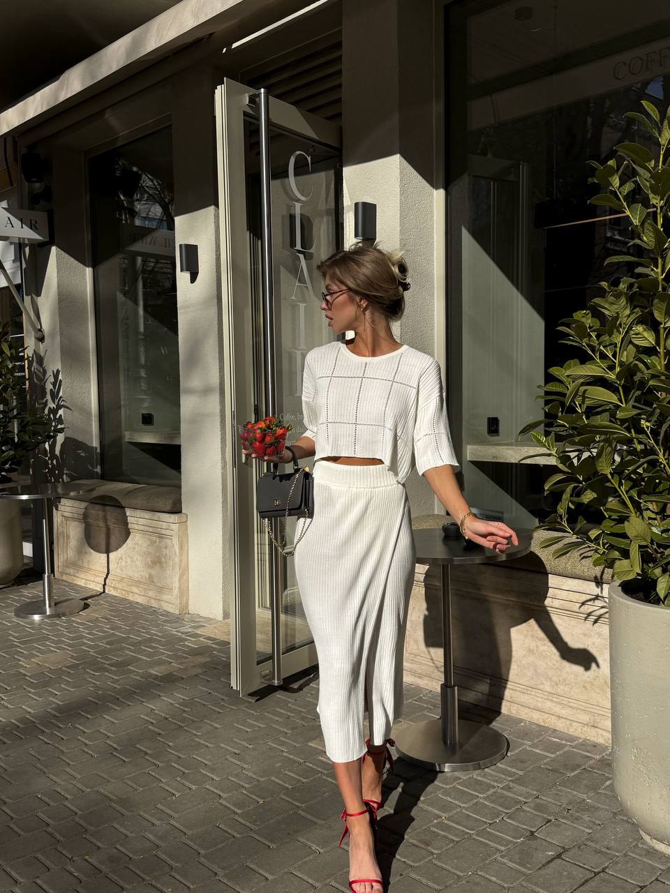 Woman in a white outfit standing outside a modern building with plants around.