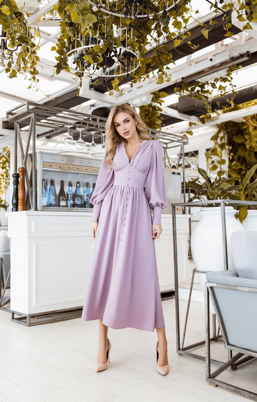 Woman in a lavender dress standing in an outdoor setting with plants and a bar.
