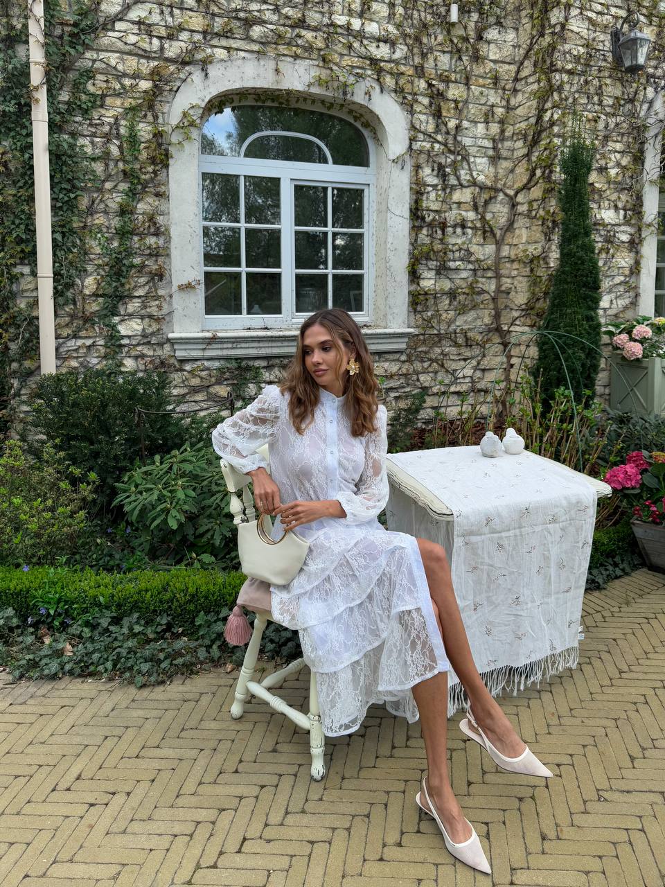 Woman in a white lace dress sitting on a chair outdoors with a stone building and garden in the background.