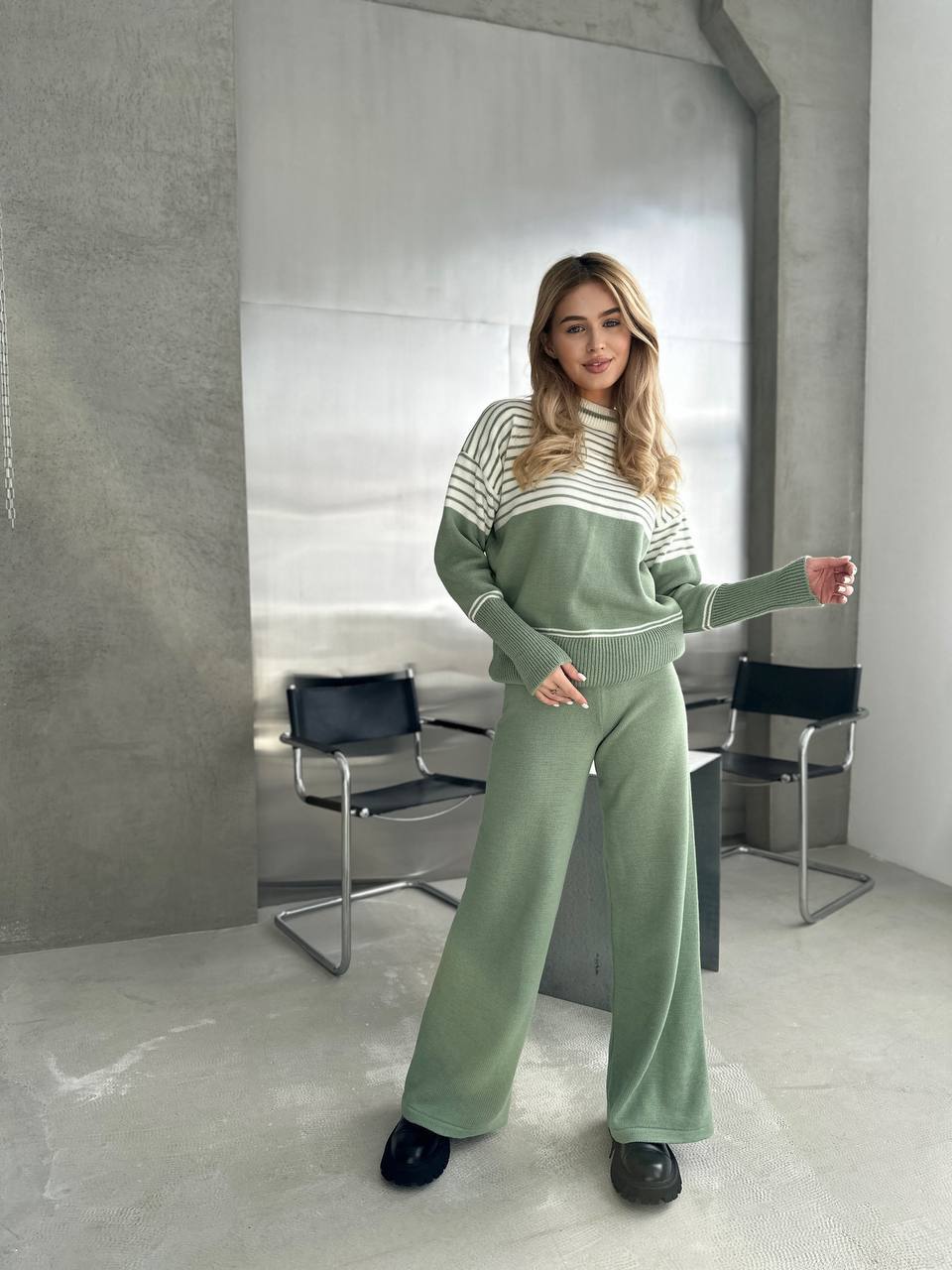Woman in a green tracksuit standing in a modern room with concrete walls and black chairs.