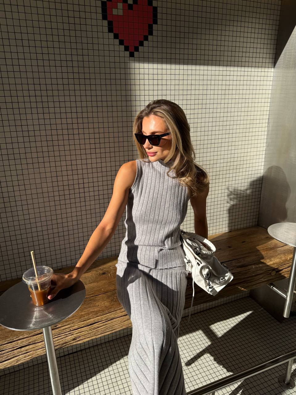 Woman in a gray outfit sitting at a table with a drink, wearing sunglasses.