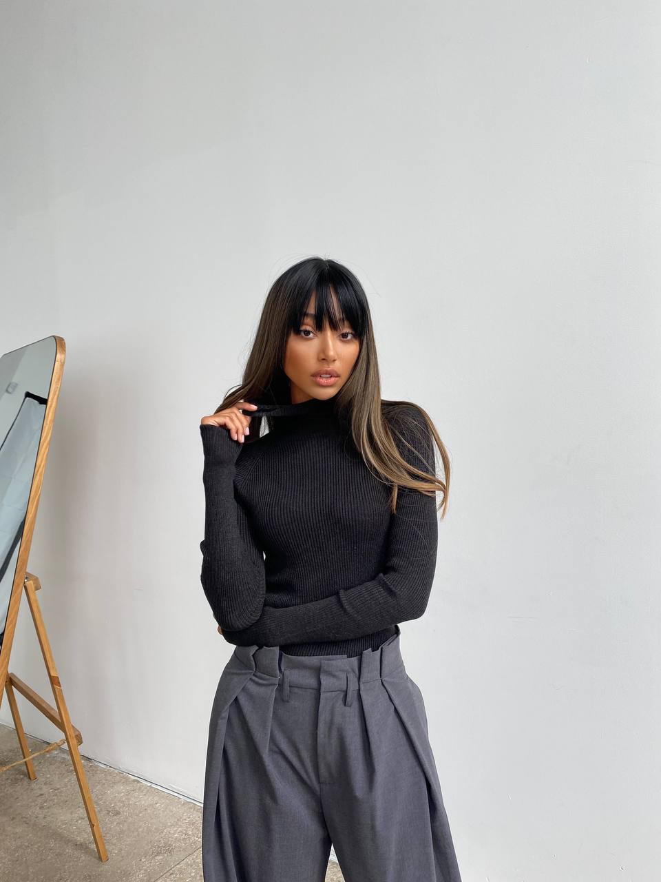 Woman wearing a black sweater and gray pants against a white wall