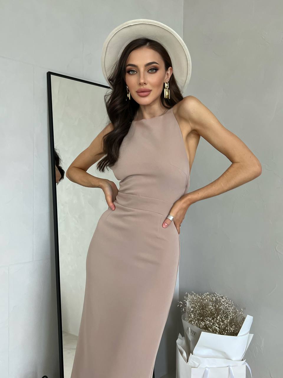 Woman in a beige dress and white hat standing in front of a mirror.