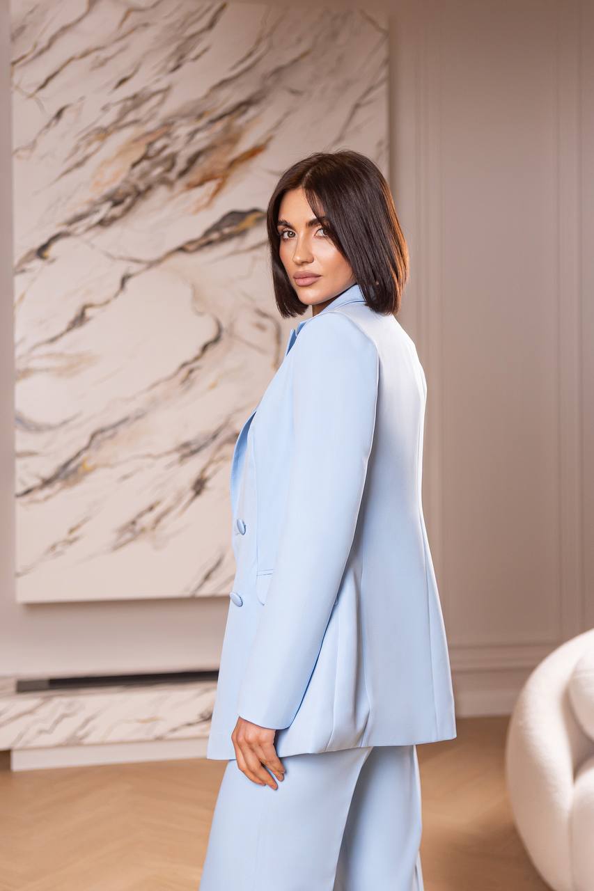 Woman in a light blue suit standing in a room with marble wall and white chair.