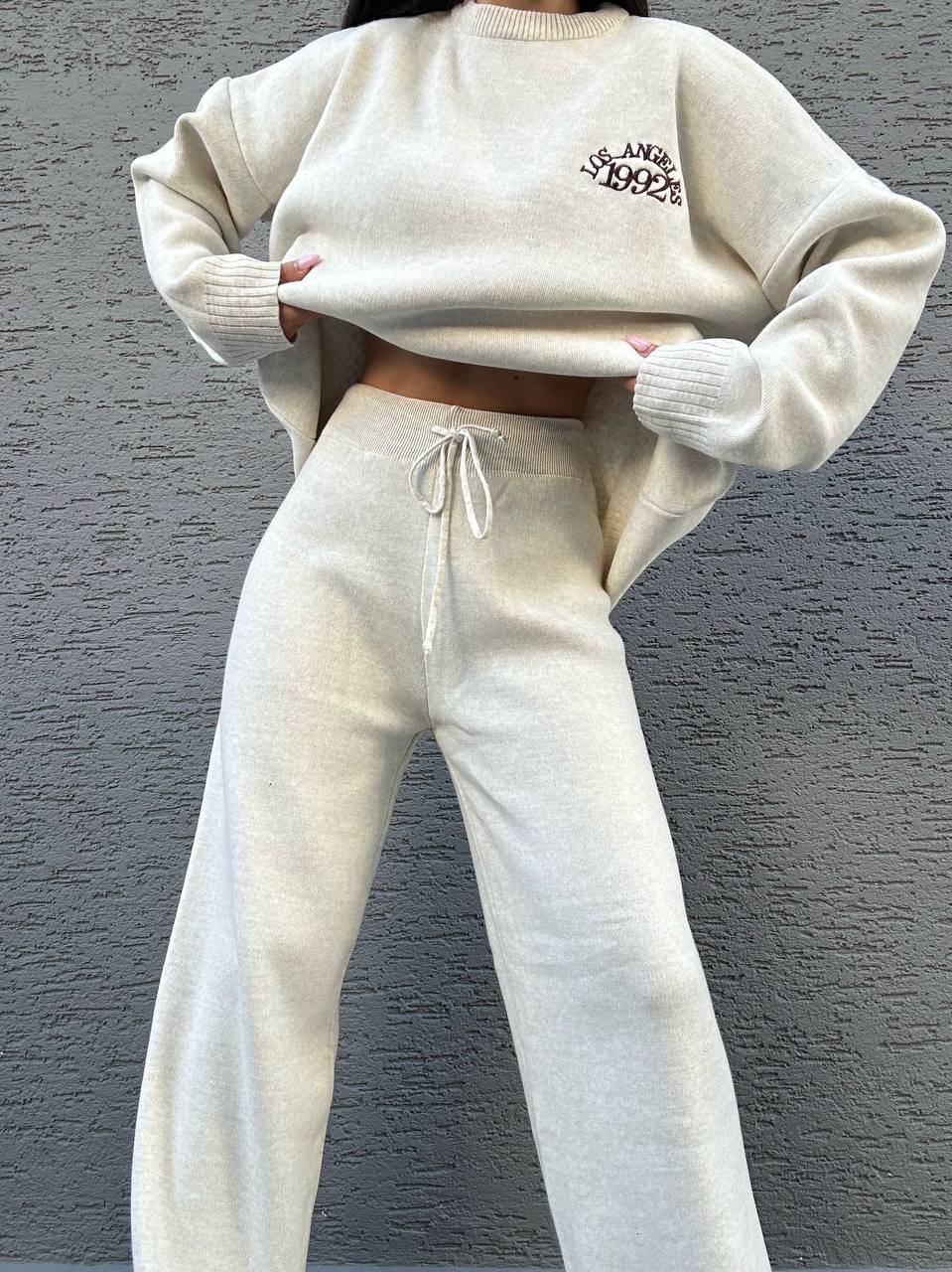 Person wearing a beige sweater and matching pants against a gray background