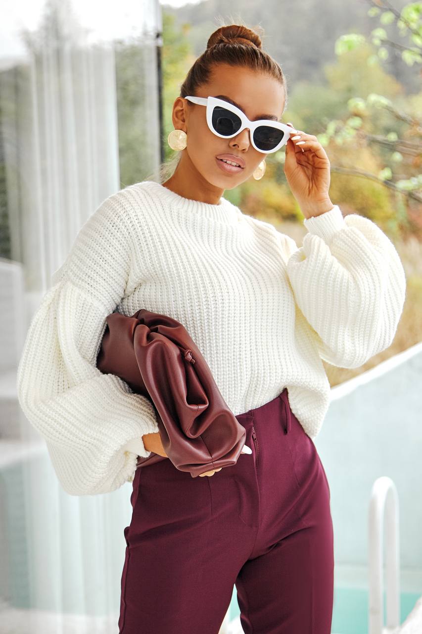 Woman wearing a white sweater and burgundy pants, holding a brown jacket, outdoors.