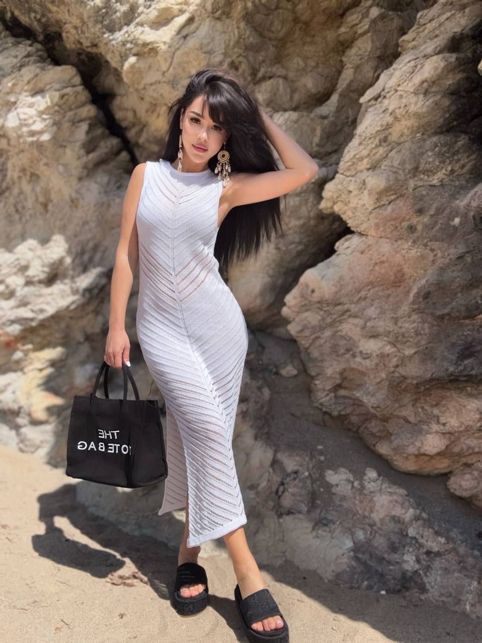 Woman in a white dress holding a black handbag against a rocky background