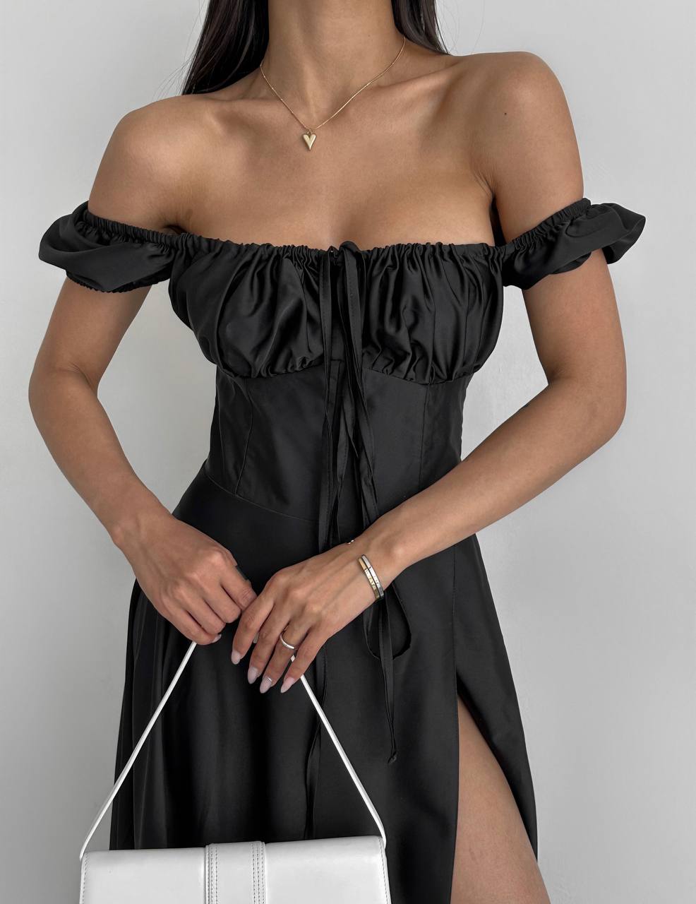 Woman wearing a black off-shoulder dress with a white handbag on a plain background