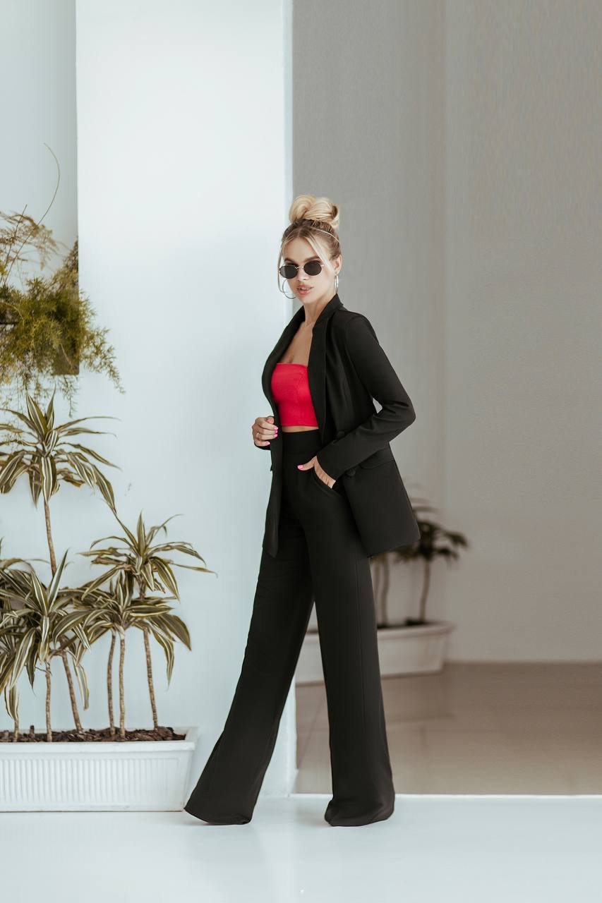 Woman in a black outfit with a red top standing indoors against a white wall.