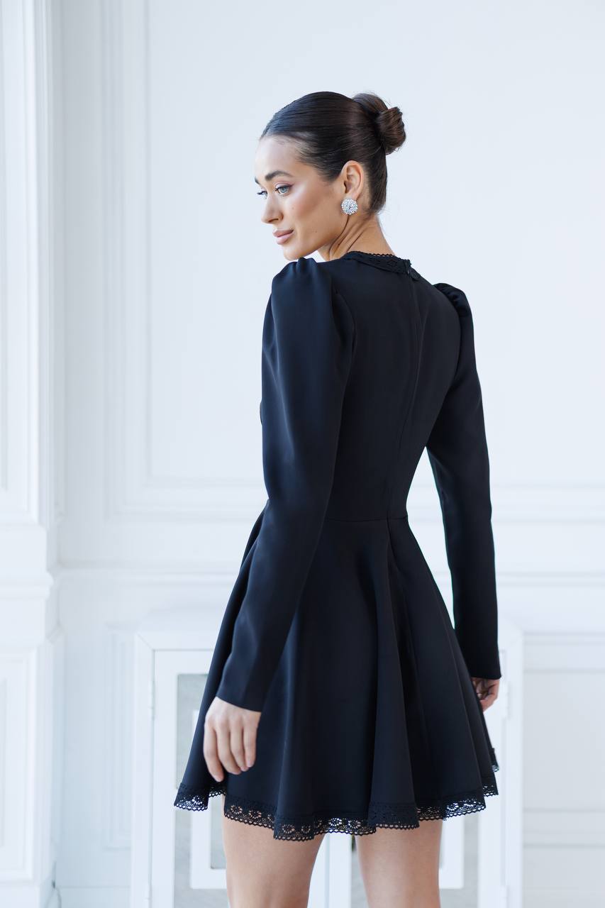 Woman wearing a black dress with long sleeves and lace trim in a minimalistic setting.