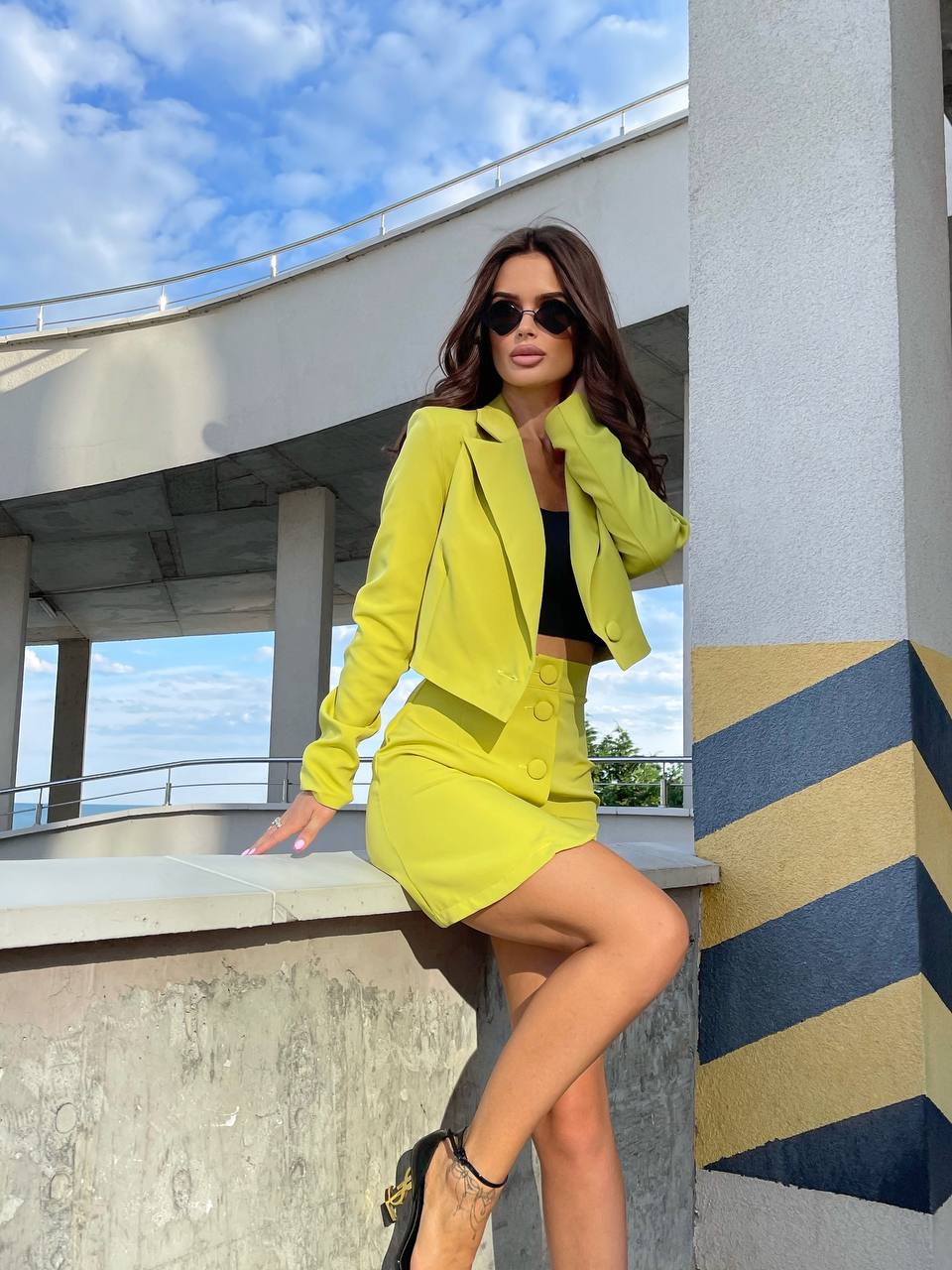 Woman in a bright yellow outfit posing outdoors with a blue sky and modern architecture.