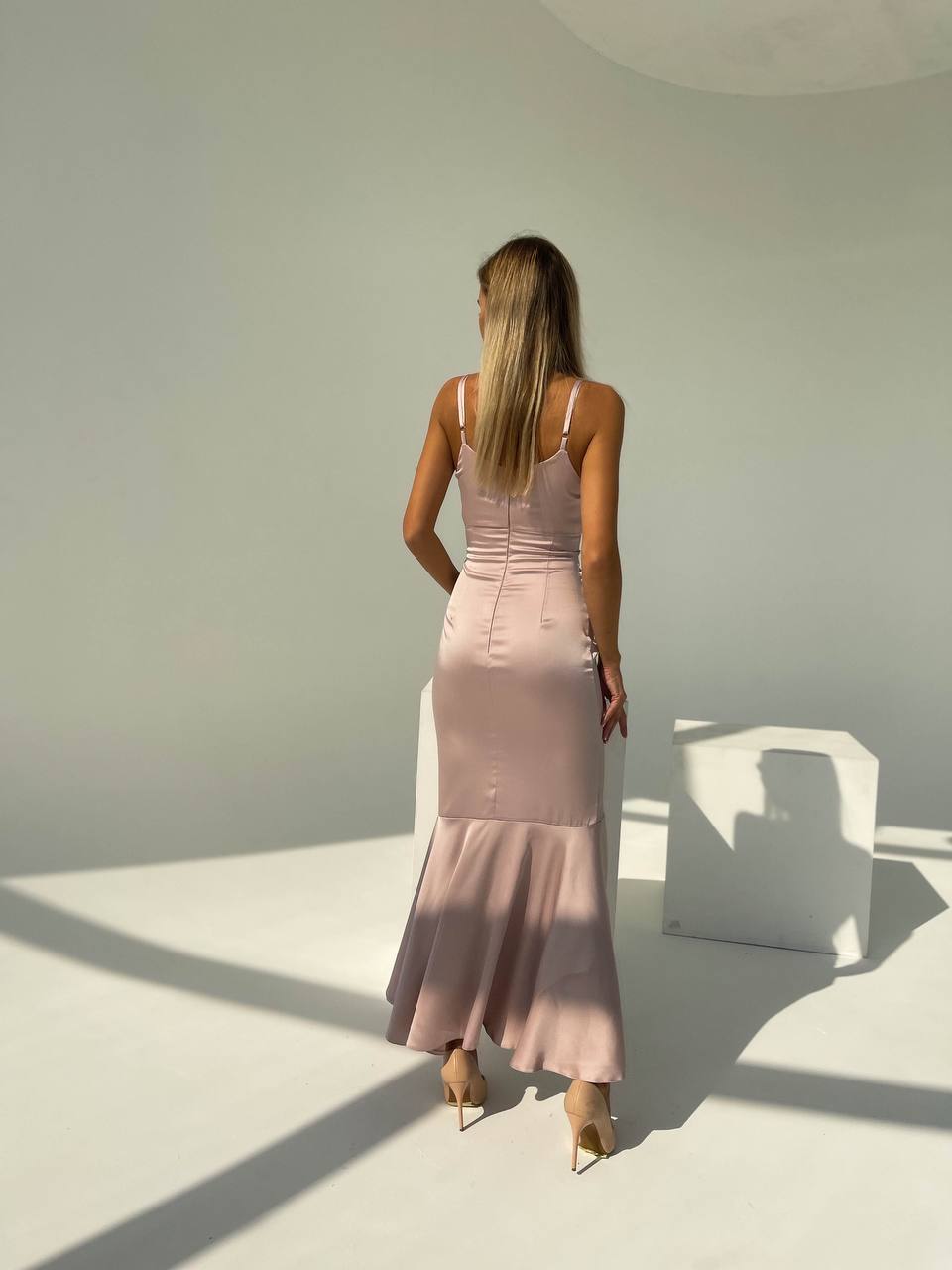 Woman in a pink dress standing in a minimalistic room with sunlight casting shadows.