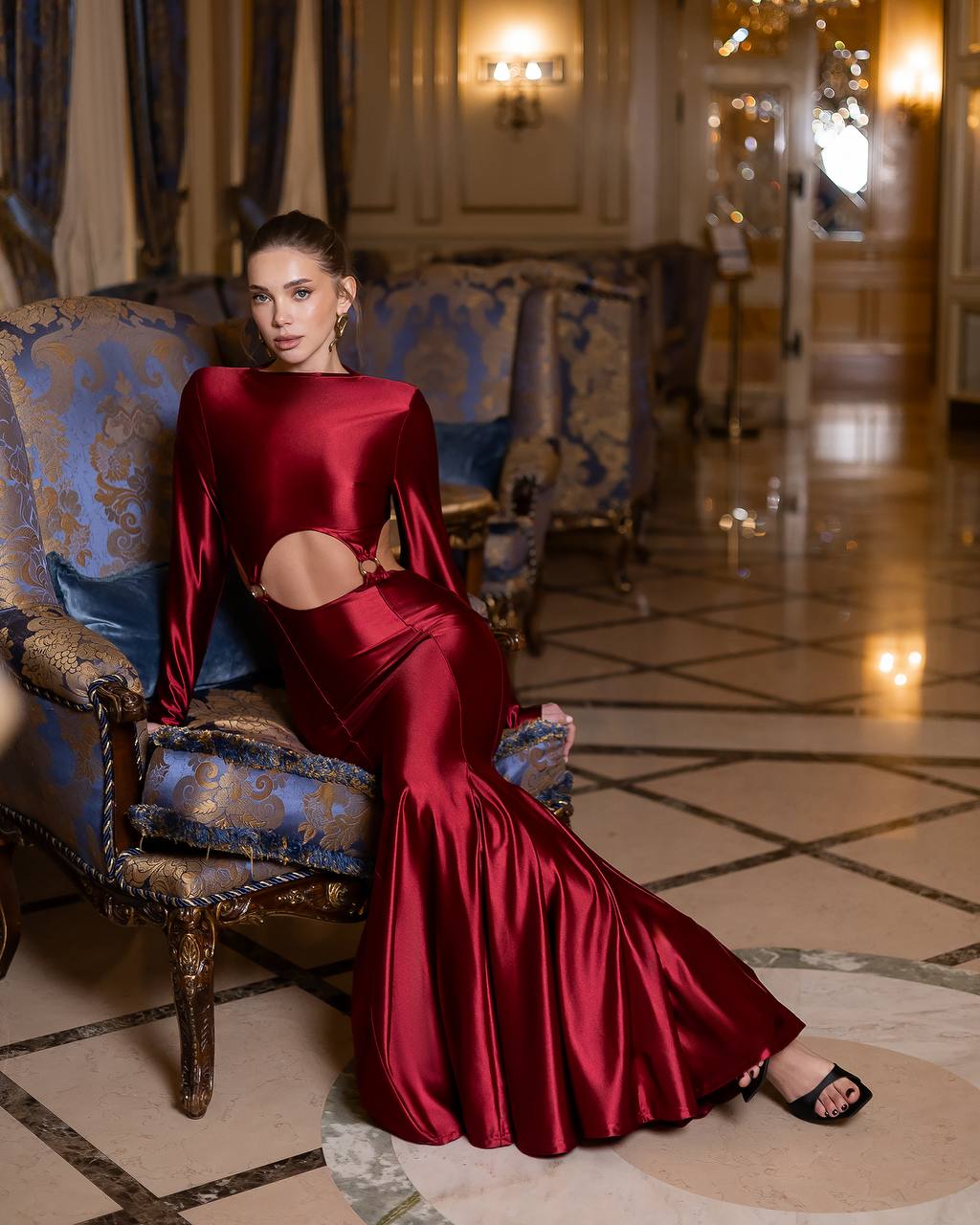Woman in a red dress sitting on an ornate chair in a luxurious room.