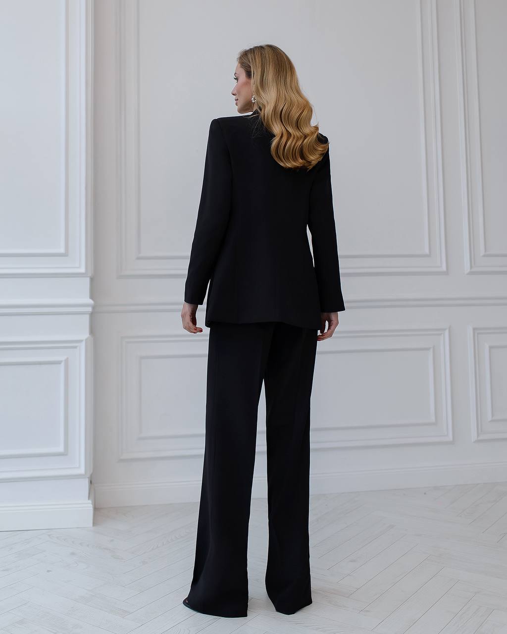 Woman wearing a black suit standing against a white paneled wall.