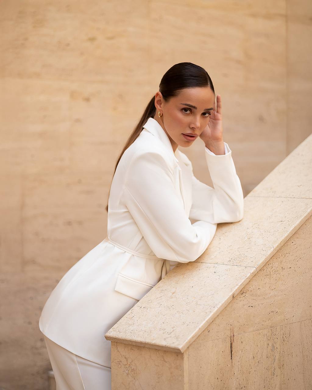 Woman in a white outfit sitting on a stone ledge against a beige wall.