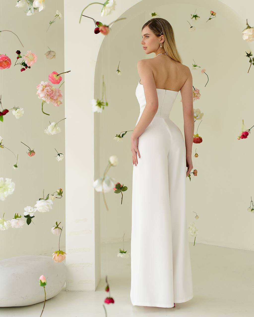 Woman in a white strapless jumpsuit standing in a room with floral decorations.