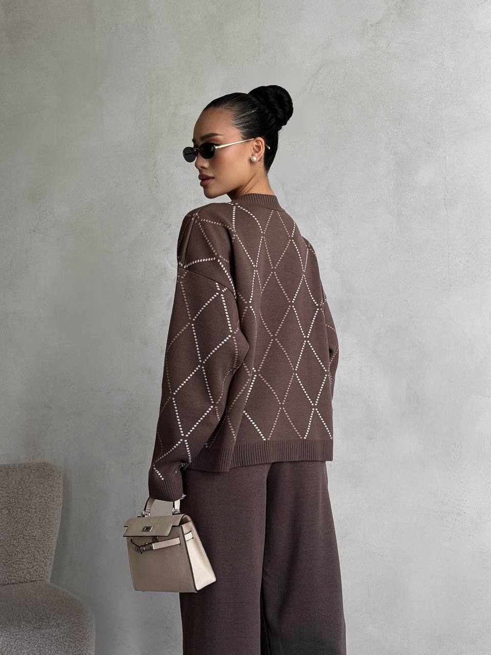 Woman wearing a brown patterned sweater and pants, holding a beige handbag against a gray background.
