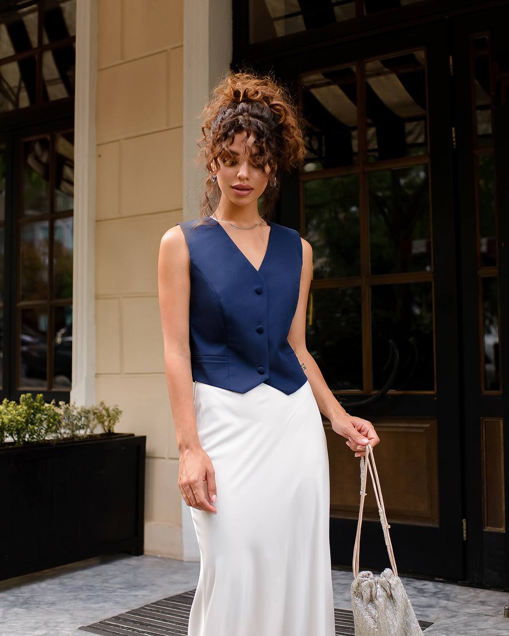 Woman wearing a blue sleeveless top and white skirt standing outside a building.