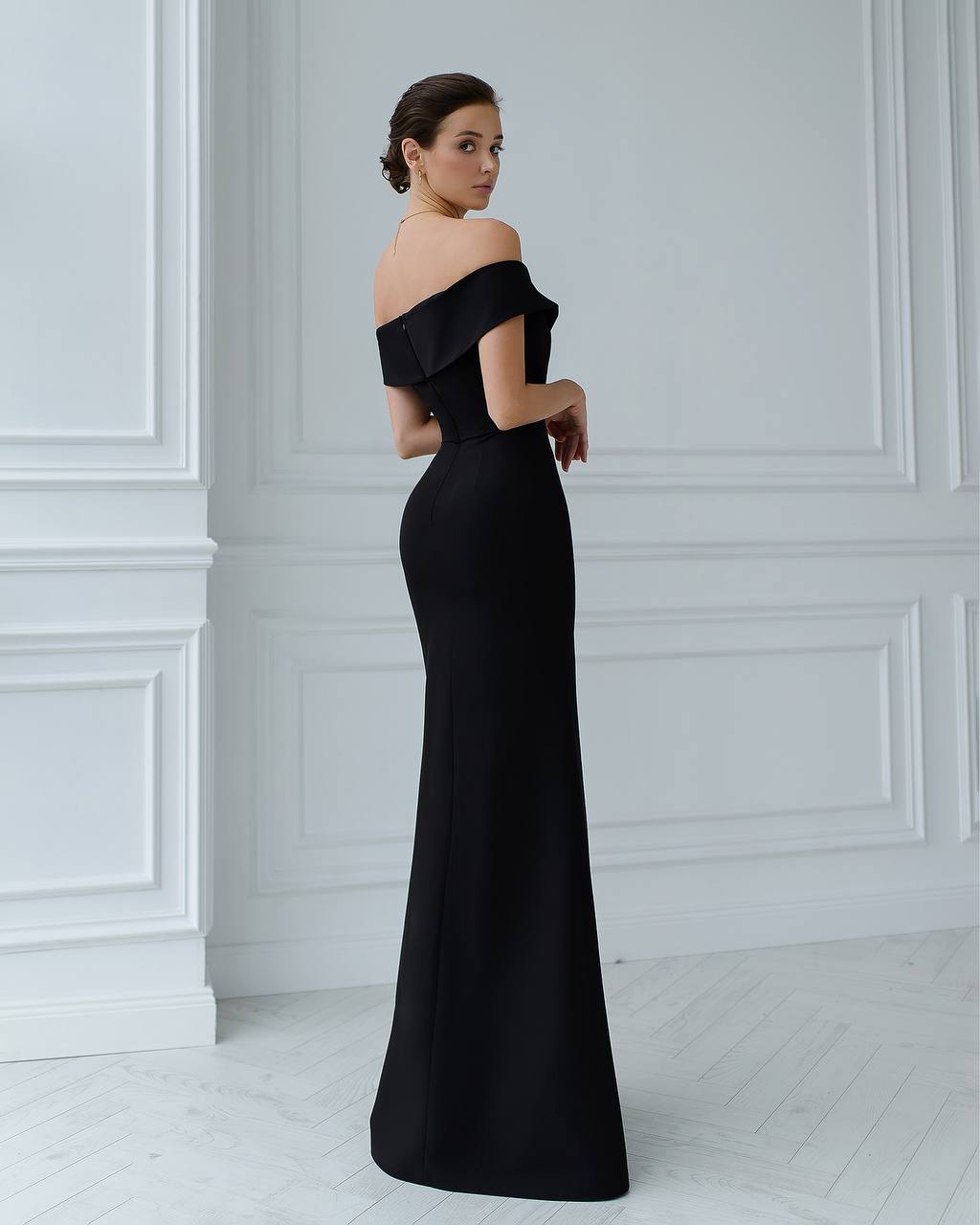 Woman wearing a black off-shoulder evening gown against a white wall.