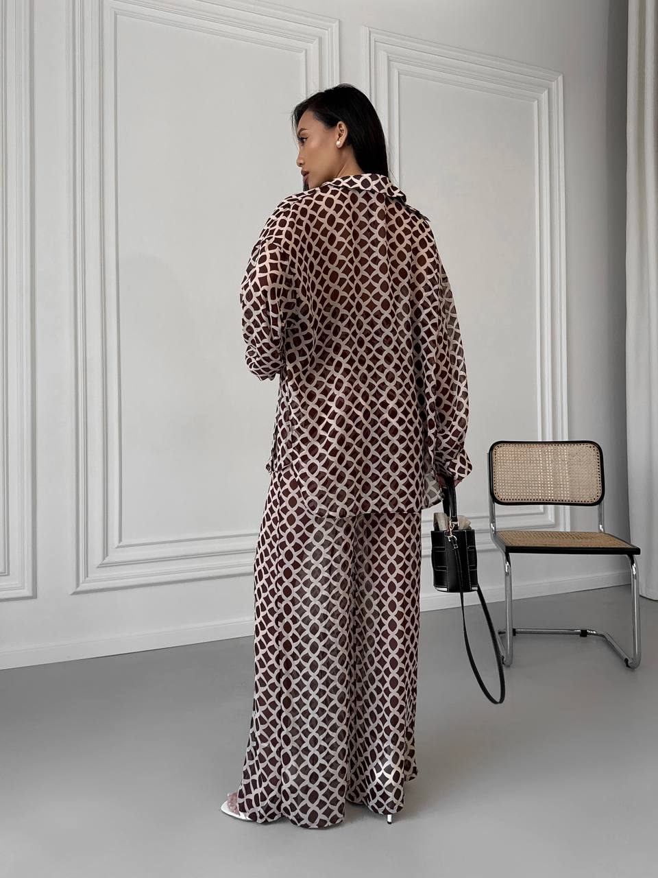 Woman in a patterned outfit standing in a room with a chair and wall.