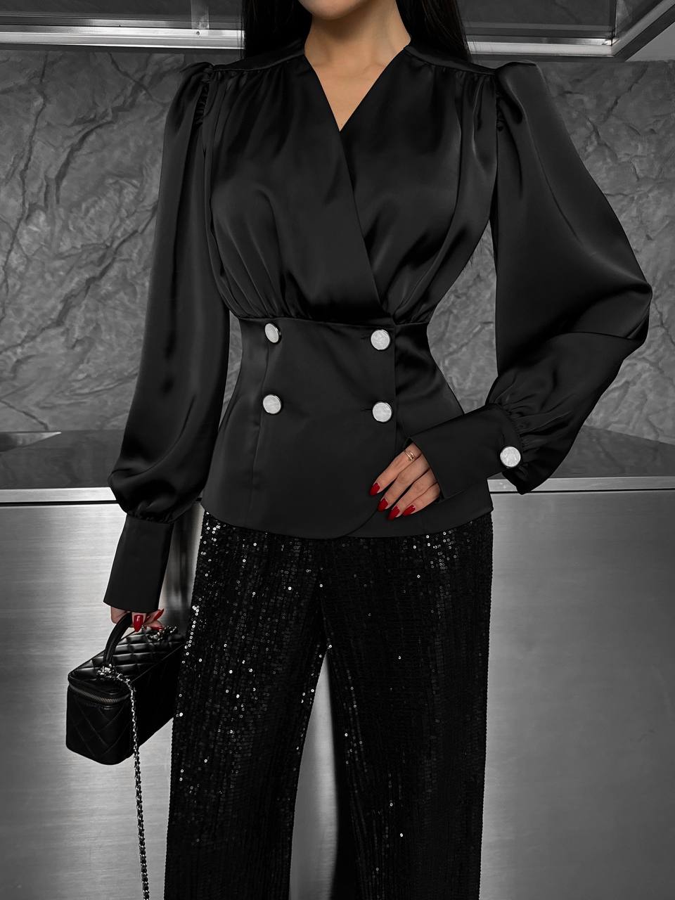 Person wearing a black blouse with white buttons and black sequin pants, holding a black handbag.